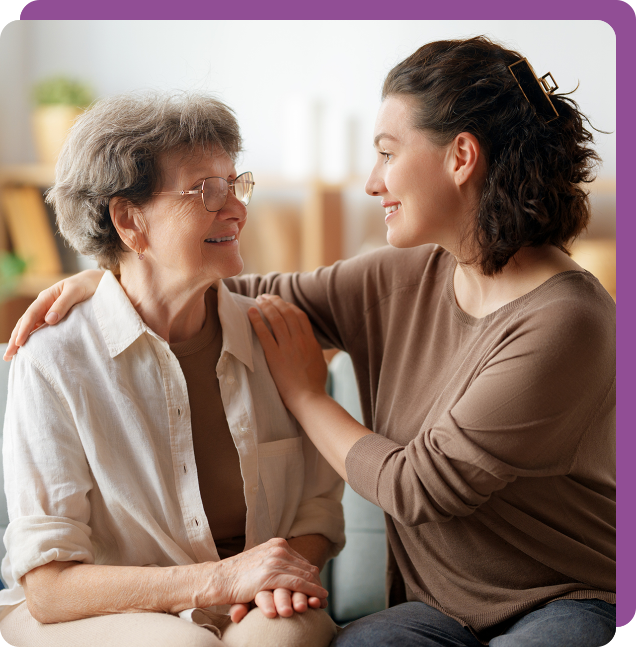 A young woman warmly supporting an elderly woman in a cozy setting.