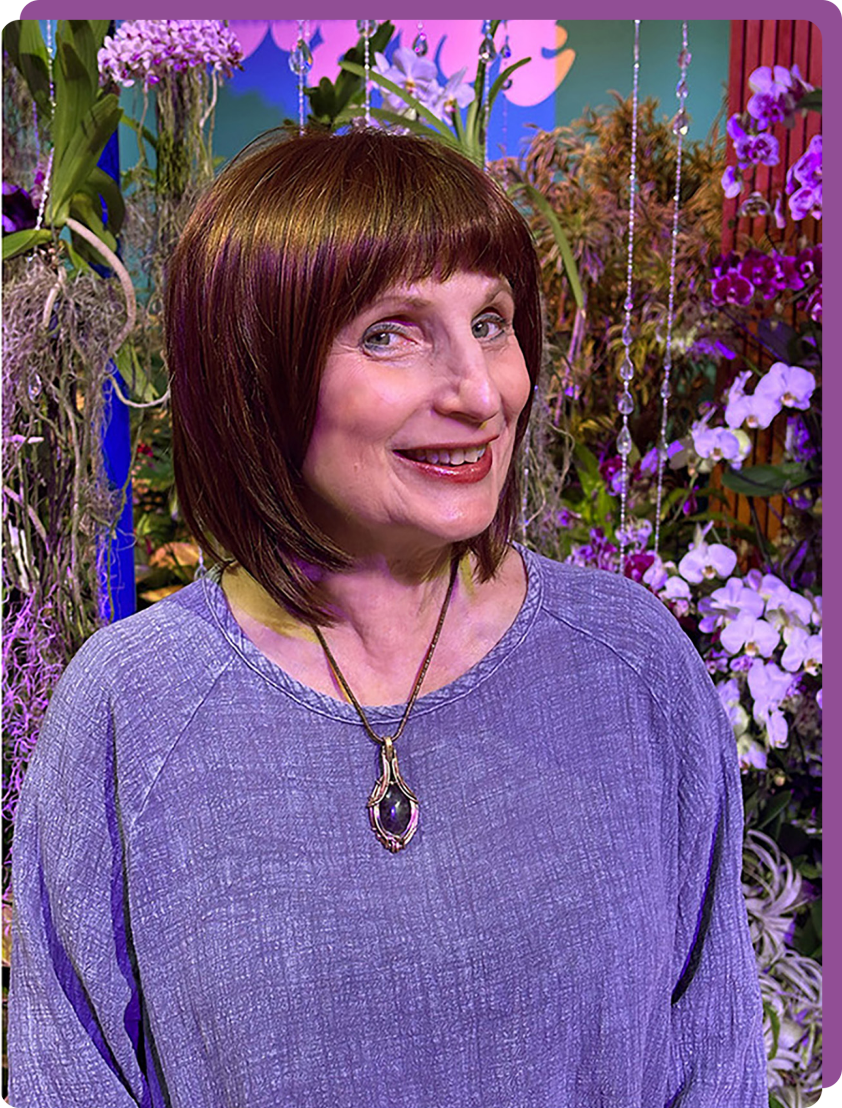 Smiling woman in a purple top with a pendant necklace, standing among vibrant flowers.