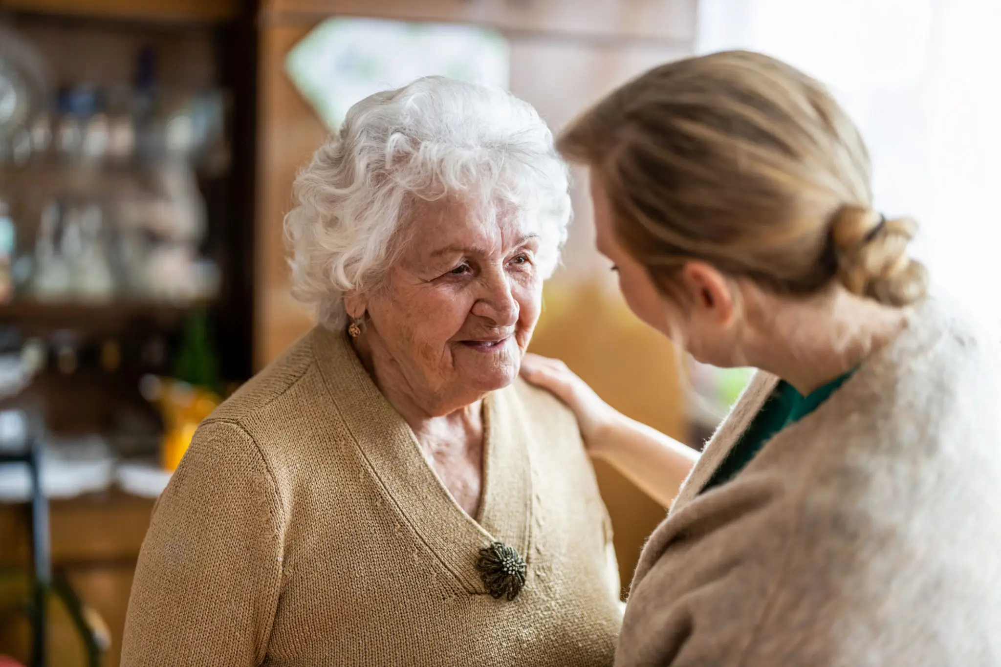 A caregiver warmly interacts with an elderly woman in a cozy home setting.